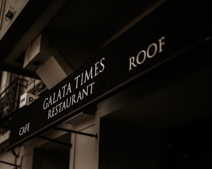 A black signboard features the text 'Galata Times Restaurant' along with 'Cafe' and 'Roof,' displayed in white letters. The scene includes the building's architectural elements with a slightly angled perspective, and the signage is under a dim, warm lighting that creates a vintage atmosphere.