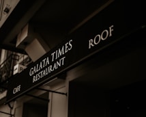 A black signboard features the text 'Galata Times Restaurant' along with 'Cafe' and 'Roof,' displayed in white letters. The scene includes the building's architectural elements with a slightly angled perspective, and the signage is under a dim, warm lighting that creates a vintage atmosphere.