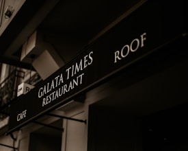 A black signboard features the text 'Galata Times Restaurant' along with 'Cafe' and 'Roof,' displayed in white letters. The scene includes the building's architectural elements with a slightly angled perspective, and the signage is under a dim, warm lighting that creates a vintage atmosphere.