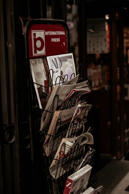A wire rack filled with various magazines and newspapers is positioned next to a door with a visible sign that reads 'International Press' on the top. The rack contains publications such as 'Architectural Digest,' 'Cornucopia,' and 'Living with Style,' among others.