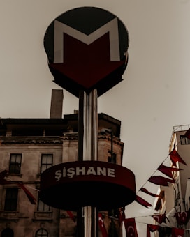 A metro sign with the word 'Şişhane' and an arrow pointing downward. The background features an urban setting with buildings and red flags bearing white symbols, likely the Turkish flag. The sky appears overcast, contributing to the overall atmosphere.