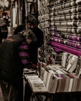 Two people are closely examining books and postcards at a small outdoor stall. The setting appears to be a narrow street or alley lined with merchandise. The background shows a partially closed, graffiti-covered shutter, and one of the individuals wears a jacket with branding on the back.