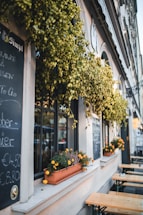 A quaint outdoor cafe with chalkboards displaying menu items. Lush green vines hang above the window, and bright orange flowers sit in window boxes. Wooden picnic tables are arranged outside on the street, offering a cozy and inviting atmosphere.