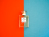 A perfume bottle with a clear, rectangular design is positioned against a background divided into bright orange on the left and vivid blue on the right. The label on the bottle reads 'N°5 CHANEL PARIS EAU DE PARFUM'. The bottle cap is transparent and matches the sophisticated and elegant aesthetic of the fragrance.