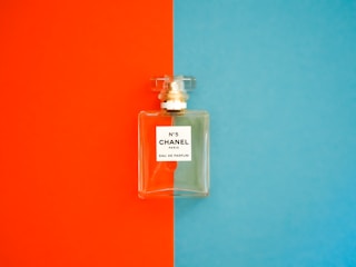 A perfume bottle with a clear, rectangular design is positioned against a background divided into bright orange on the left and vivid blue on the right. The label on the bottle reads 'N°5 CHANEL PARIS EAU DE PARFUM'. The bottle cap is transparent and matches the sophisticated and elegant aesthetic of the fragrance.
