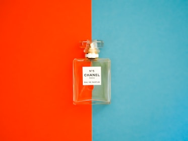 A perfume bottle with a clear, rectangular design is positioned against a background divided into bright orange on the left and vivid blue on the right. The label on the bottle reads 'N°5 CHANEL PARIS EAU DE PARFUM'. The bottle cap is transparent and matches the sophisticated and elegant aesthetic of the fragrance.