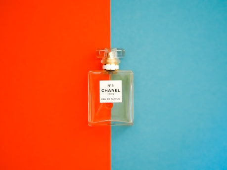 A perfume bottle with a clear, rectangular design is positioned against a background divided into bright orange on the left and vivid blue on the right. The label on the bottle reads 'N°5 CHANEL PARIS EAU DE PARFUM'. The bottle cap is transparent and matches the sophisticated and elegant aesthetic of the fragrance.