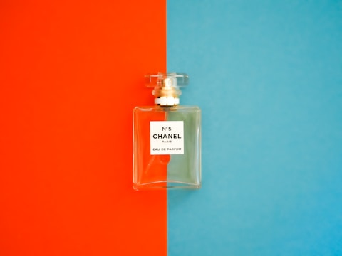 A perfume bottle with a clear, rectangular design is positioned against a background divided into bright orange on the left and vivid blue on the right. The label on the bottle reads 'N°5 CHANEL PARIS EAU DE PARFUM'. The bottle cap is transparent and matches the sophisticated and elegant aesthetic of the fragrance.