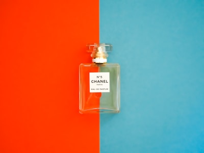 A perfume bottle with a clear, rectangular design is positioned against a background divided into bright orange on the left and vivid blue on the right. The label on the bottle reads 'N°5 CHANEL PARIS EAU DE PARFUM'. The bottle cap is transparent and matches the sophisticated and elegant aesthetic of the fragrance.