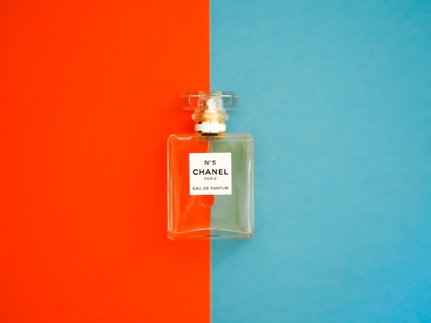 A perfume bottle with a clear, rectangular design is positioned against a background divided into bright orange on the left and vivid blue on the right. The label on the bottle reads 'N°5 CHANEL PARIS EAU DE PARFUM'. The bottle cap is transparent and matches the sophisticated and elegant aesthetic of the fragrance.