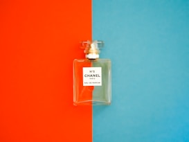 A perfume bottle with a clear, rectangular design is positioned against a background divided into bright orange on the left and vivid blue on the right. The label on the bottle reads 'N°5 CHANEL PARIS EAU DE PARFUM'. The bottle cap is transparent and matches the sophisticated and elegant aesthetic of the fragrance.