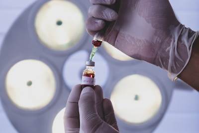 A person wearing gloves is handling a vial labeled 'Covid-19' with a syringe, possibly extracting or injecting fluid. The background shows out-of-focus circular lights, likely from medical equipment or a laboratory setting.