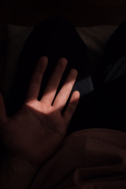 A hand is positioned palm up, illuminated by a soft, focused beam of light. Surrounding the hand is a dark background, which creates a stark contrast and emphasizes the lighting on the palm.