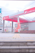 A car rental facility with noticeable signage in red and white is in view. The entrance shows a red vending machine and two trash bins nearby. The surroundings include buildings with visible architectural elements, and the area has some stairs leading up to the rental space.