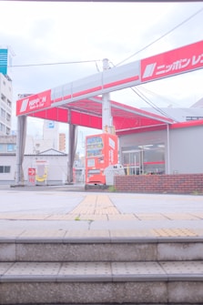 A car rental facility with noticeable signage in red and white is in view. The entrance shows a red vending machine and two trash bins nearby. The surroundings include buildings with visible architectural elements, and the area has some stairs leading up to the rental space.