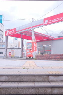 A car rental facility with noticeable signage in red and white is in view. The entrance shows a red vending machine and two trash bins nearby. The surroundings include buildings with visible architectural elements, and the area has some stairs leading up to the rental space.
