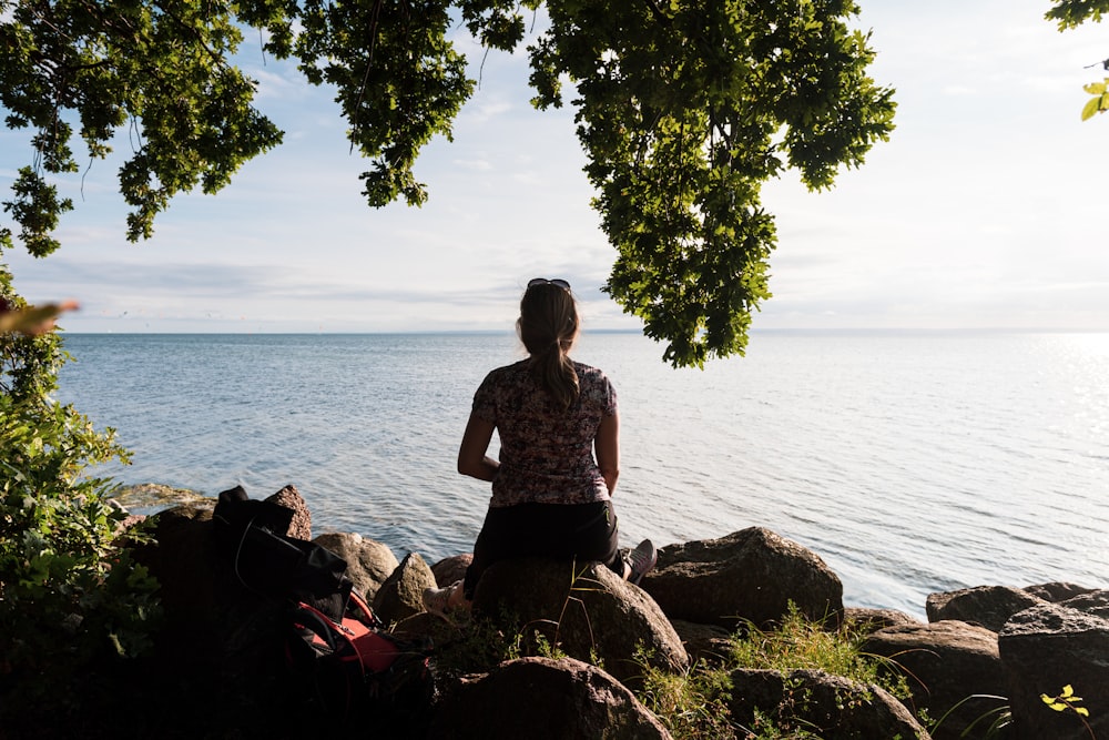 woman in black and white floral dress sitting on brown rock near body of water during