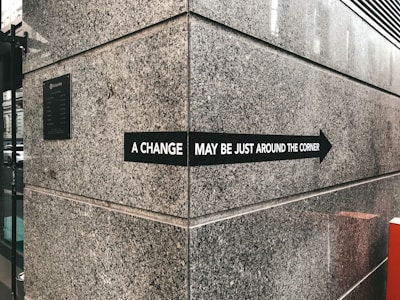 A stone wall with a black sign that has white text reads 'A change may be just around the corner' with an arrow pointing to the right. The wall is part of a corner section of a building, with visible textures of large granite-like panels. A plaque is affixed to the wall to the left, and part of a cityscape with buildings and cars can be seen in the reflection of a window on the left side.