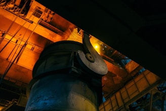 An industrial setting with large metal structures and containers, bathed in a warm orange light. The image captures machinery and equipment, possibly in a factory or manufacturing plant, with contrasting shadows and highlights.