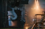 A large industrial setting with heavy machinery and metal structures. The environment is dimly lit with warm tones from the lights above, casting a dramatic glow and shadows. The focus is on a sizable metal container suspended from a crane, situated in an expansive, high-ceilinged factory space. Staircases and railings are seen to the right, adding depth to the scene.