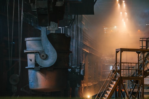A large industrial setting with heavy machinery and metal structures. The environment is dimly lit with warm tones from the lights above, casting a dramatic glow and shadows. The focus is on a sizable metal container suspended from a crane, situated in an expansive, high-ceilinged factory space. Staircases and railings are seen to the right, adding depth to the scene.