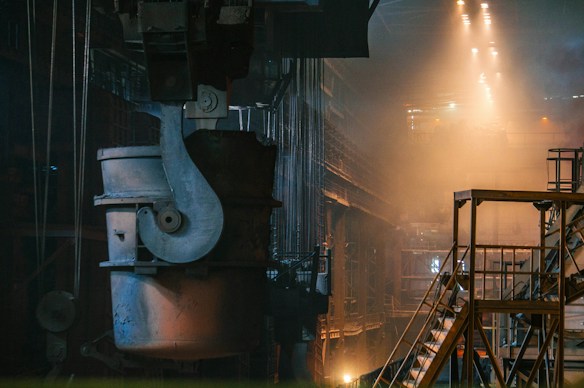A large industrial setting with heavy machinery and metal structures. The environment is dimly lit with warm tones from the lights above, casting a dramatic glow and shadows. The focus is on a sizable metal container suspended from a crane, situated in an expansive, high-ceilinged factory space. Staircases and railings are seen to the right, adding depth to the scene.