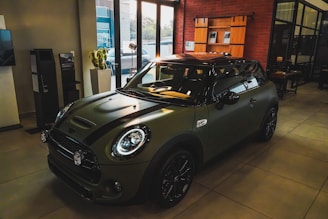 A sleek, modern Mini Cooper is parked inside a well-lit showroom with large windows. The car has a matte dark green finish with black racing stripes and polished black wheels. Various pieces of modern furniture and a potted plant can be seen in the background.