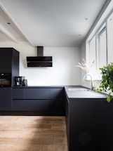 A modern kitchen with sleek black cabinets and countertops contrasts with light wooden flooring. A stainless steel faucet is set into the counter near the window, where a small plant with green leaves rests. A black range hood is mounted against a white wall, and a coffee maker with a cup is visible on the countertop.