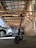 An electric scooter is parked inside a spacious garage with a slanted wooden roof and exposed brick walls. Several cars are also parked inside, and the lighting is provided by natural light coming through the openings.