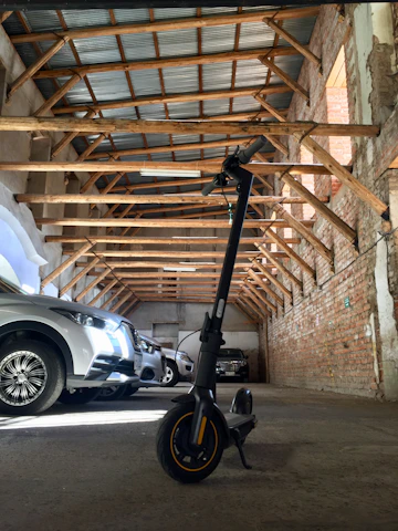 An electric scooter is parked inside a spacious garage with a slanted wooden roof and exposed brick walls. Several cars are also parked inside, and the lighting is provided by natural light coming through the openings.