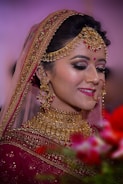 A bride adorned in elaborate gold jewelry, including a necklace, earrings, and headpiece, complemented by a red bindi. She wears a richly embroidered maroon saree with a veil edged in gold embroidery. Her makeup is elegant, with dark eye shadow and bright pink lipstick. The background is softly blurred with hints of green and red from floral arrangements in the foreground.