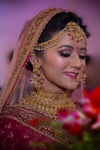 A bride adorned in elaborate gold jewelry, including a necklace, earrings, and headpiece, complemented by a red bindi. She wears a richly embroidered maroon saree with a veil edged in gold embroidery. Her makeup is elegant, with dark eye shadow and bright pink lipstick. The background is softly blurred with hints of green and red from floral arrangements in the foreground.