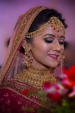 A bride adorned in elaborate gold jewelry, including a necklace, earrings, and headpiece, complemented by a red bindi. She wears a richly embroidered maroon saree with a veil edged in gold embroidery. Her makeup is elegant, with dark eye shadow and bright pink lipstick. The background is softly blurred with hints of green and red from floral arrangements in the foreground.