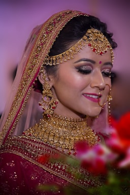 A bride adorned in elaborate gold jewelry, including a necklace, earrings, and headpiece, complemented by a red bindi. She wears a richly embroidered maroon saree with a veil edged in gold embroidery. Her makeup is elegant, with dark eye shadow and bright pink lipstick. The background is softly blurred with hints of green and red from floral arrangements in the foreground.