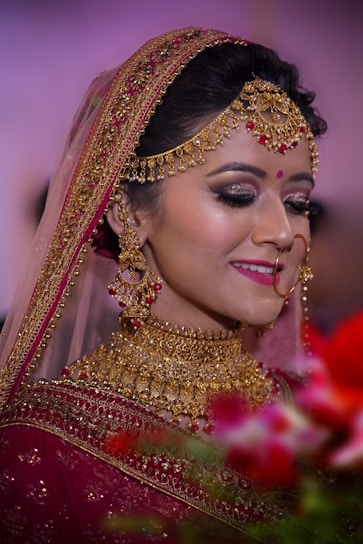 A bride adorned in elaborate gold jewelry, including a necklace, earrings, and headpiece, complemented by a red bindi. She wears a richly embroidered maroon saree with a veil edged in gold embroidery. Her makeup is elegant, with dark eye shadow and bright pink lipstick. The background is softly blurred with hints of green and red from floral arrangements in the foreground.