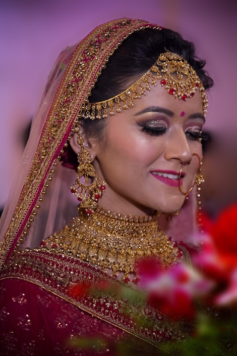 A bride adorned in elaborate gold jewelry, including a necklace, earrings, and headpiece, complemented by a red bindi. She wears a richly embroidered maroon saree with a veil edged in gold embroidery. Her makeup is elegant, with dark eye shadow and bright pink lipstick. The background is softly blurred with hints of green and red from floral arrangements in the foreground.