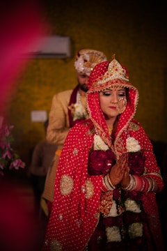 A bride in traditional red attire, adorned with intricate jewelry, stands with her eyes closed, hands pressed together in a gesture of prayer or greeting. A groom, partially visible behind her, wears golden and maroon clothing and a decorative turban.