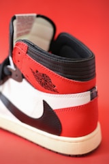 A close-up view of a sneaker, featuring a red, white, and black color scheme. The design is classic with prominent branding on the side, showcasing a logo in black against a red backdrop. The interior appears cushioned, and the sneaker maintains a high-top structure.
