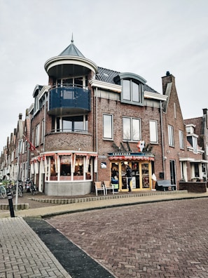A corner building features a snack bar with signage that reads 'Verse Vlaamse Frites'. The building is constructed with red bricks and has a distinct blue balcony on the upper floor. There are several large windows, a decorative gable, and a circular turret-like structure at the corner. The scene includes a few people by the entrance and multiple bicycles parked along the sidewalk. The sky is overcast, creating a subdued atmosphere.