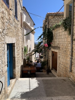 A narrow, stone-paved alleyway lined with rustic stone buildings, featuring wooden doors and blue shutters. Greenery and pink flowers climb the walls, adding charm to the scene. Outdoor tables and chairs are set up on one side, suggesting a small café or restaurant. Bright blue sky and the sea are visible in the background, giving the alley a coastal feel.