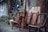 An assortment of wooden and rattan chairs is stacked and arranged against an old, rustic wall. The scene includes a folded table and various household items leaned haphazardly. The background reveals a weathered building exterior with some overgrown plants.