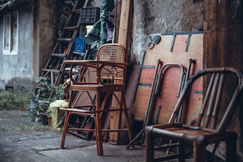 An assortment of wooden and rattan chairs is stacked and arranged against an old, rustic wall. The scene includes a folded table and various household items leaned haphazardly. The background reveals a weathered building exterior with some overgrown plants.