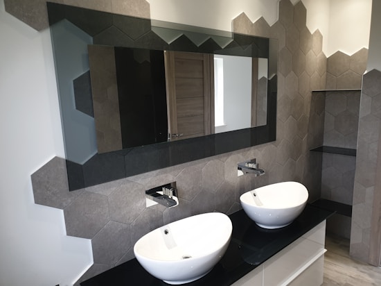 A contemporary bathroom features two modern white vessel sinks with sleek metallic faucets mounted on a glossy black countertop. The wall behind the sinks is adorned with hexagonal tiles in shades of gray and brown, creating an elegant geometric pattern. A large rectangular mirror is mounted above the sinks, reflecting part of the room, including a wooden door.