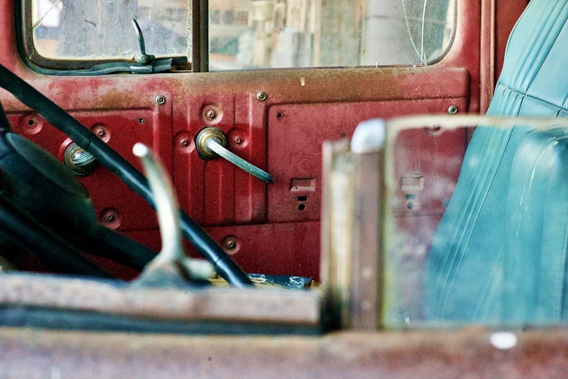 The interior of an old, rusted vehicle featuring a red metal door with a manual window crank and a blue leather seat. The space appears worn and weathered, with scratches and signs of aging on the metal and leather surfaces.