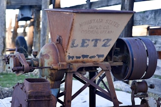 An old, rusted industrial machine with the brand name 'Letz' prominently displayed. It features gears and a large belt wheel, and is marked with the text 'Mountain States Imp. Co., Ogden, Utah'. The background shows a wooden fence and some large wheels, suggesting it might be located in a rural or farm setting.