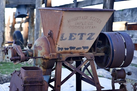 An old, rusted industrial machine with the brand name 'Letz' prominently displayed. It features gears and a large belt wheel, and is marked with the text 'Mountain States Imp. Co., Ogden, Utah'. The background shows a wooden fence and some large wheels, suggesting it might be located in a rural or farm setting.