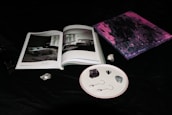 An open book with black and white images of interior designs lies on a dark surface. Next to it, a small abstract painting features swirling tones of pink and purple. A few stones and two small objects resembling earrings are neatly arranged on a white speckled plate.