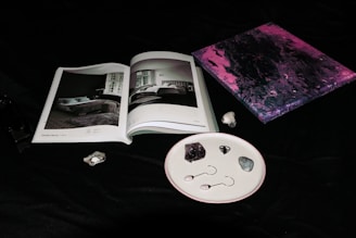 An open book with black and white images of interior designs lies on a dark surface. Next to it, a small abstract painting features swirling tones of pink and purple. A few stones and two small objects resembling earrings are neatly arranged on a white speckled plate.