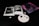 An open book with black and white images of interior designs lies on a dark surface. Next to it, a small abstract painting features swirling tones of pink and purple. A few stones and two small objects resembling earrings are neatly arranged on a white speckled plate.