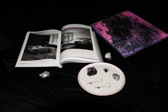 An open book with black and white images of interior designs lies on a dark surface. Next to it, a small abstract painting features swirling tones of pink and purple. A few stones and two small objects resembling earrings are neatly arranged on a white speckled plate.